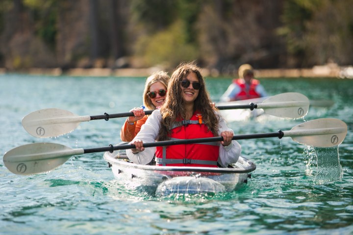 Two women kayaking on a lake with smiles, wearing red life vests.