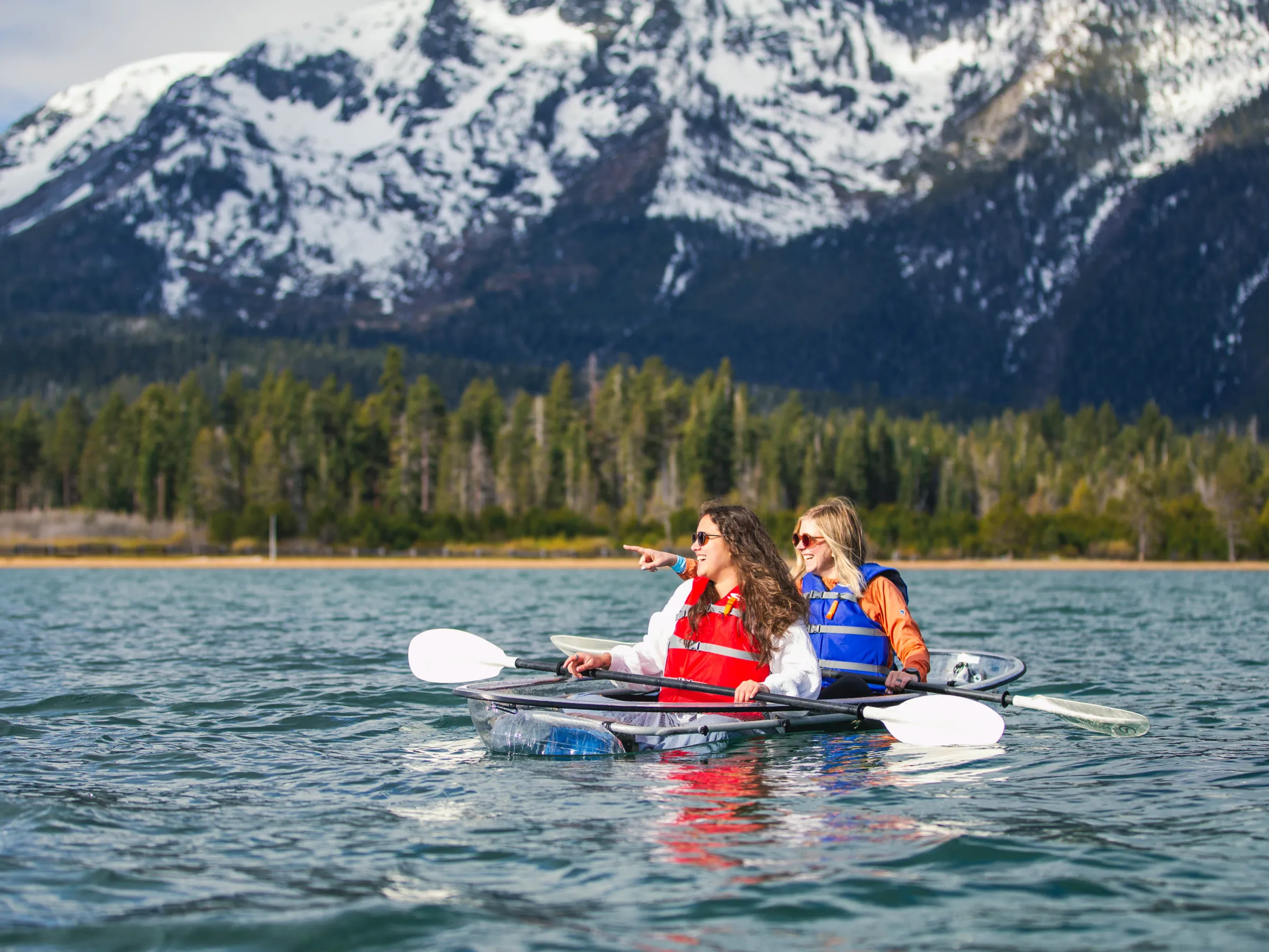 Two people kayaking on a lake with mountains and trees in the background.