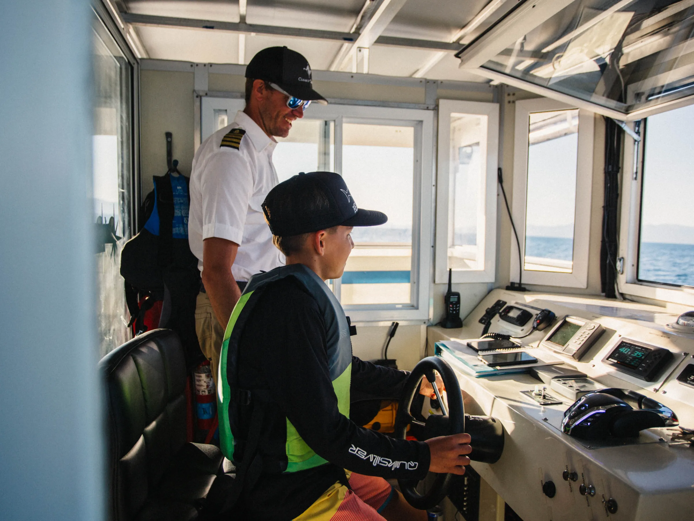 Two people in a boat cabin; one is seated at the controls, the other standing.
