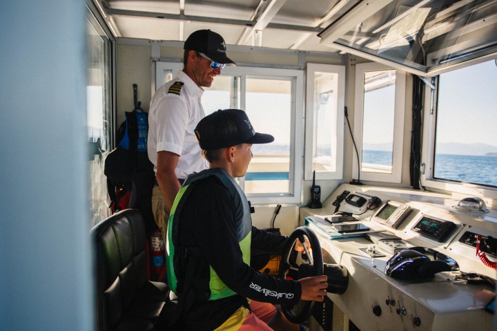 Two people in a boat cabin; one is seated at the controls, the other standing.
