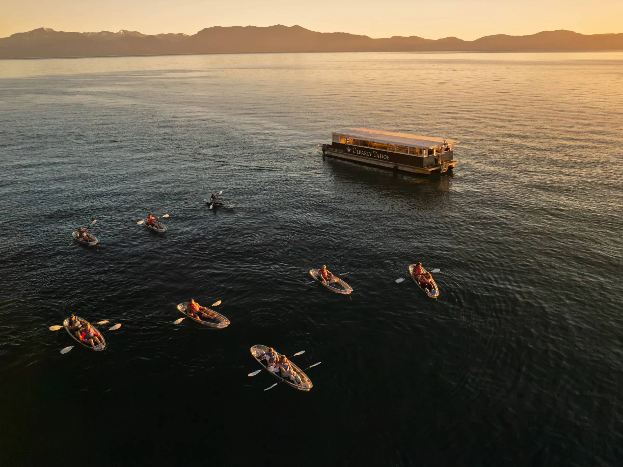 Kayakers near a large boat at sunset on a calm lake with mountains in the background.