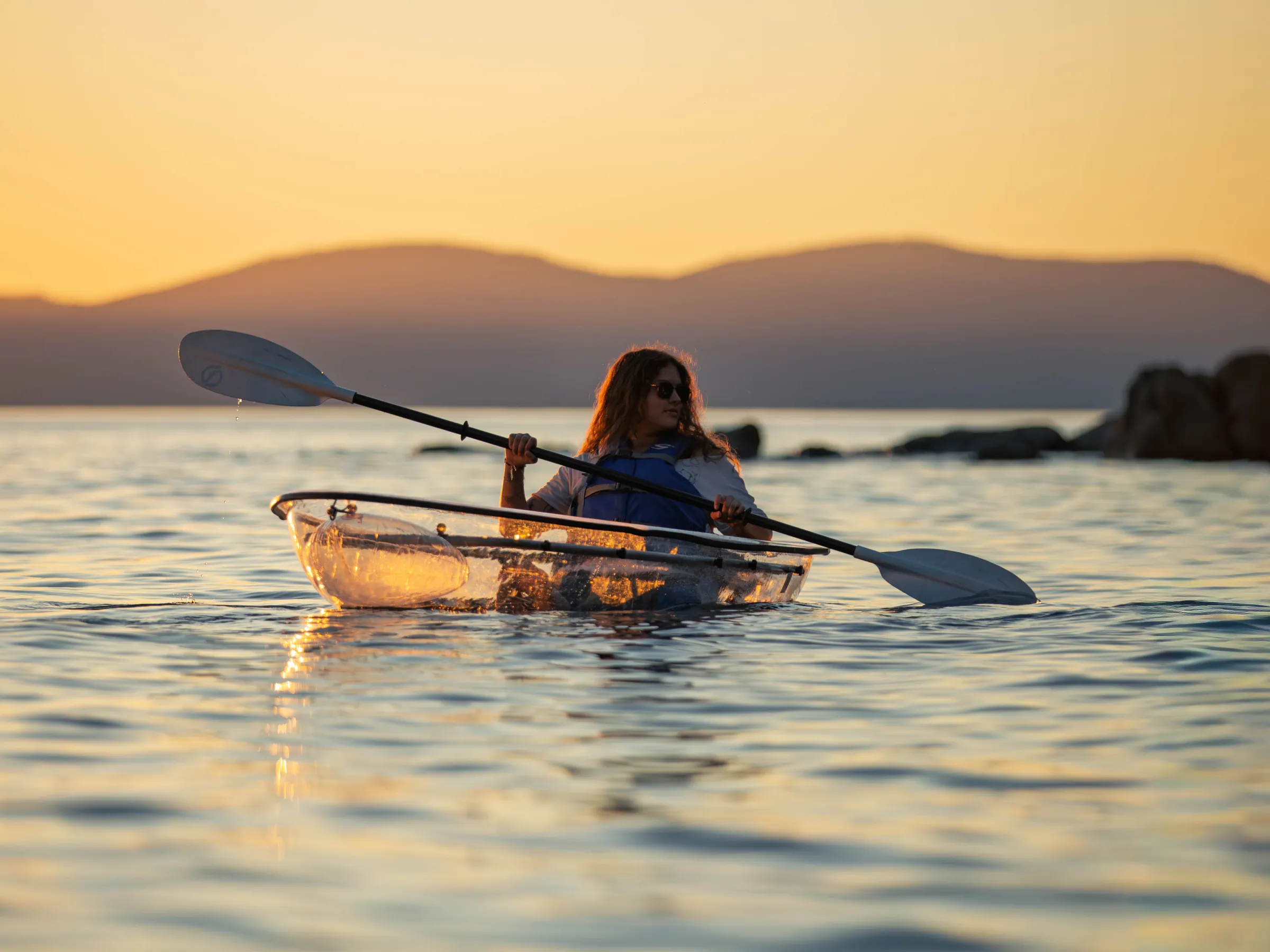 Person kayaking in transparent kayak on calm water at sunset.
