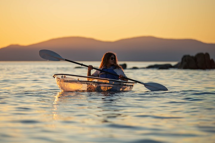Person kayaking in transparent kayak on calm water at sunset.