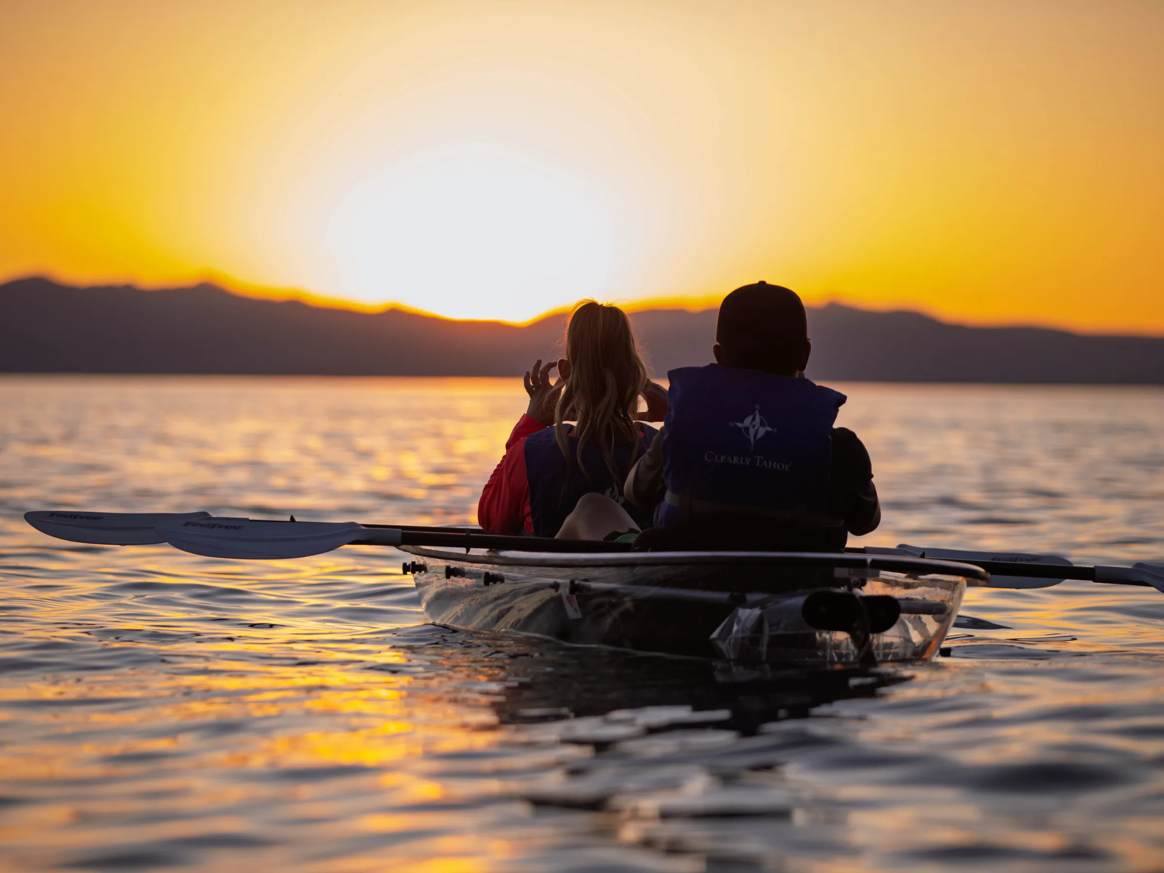 Two people kayaking on a lake at sunset with mountains in the distance.
