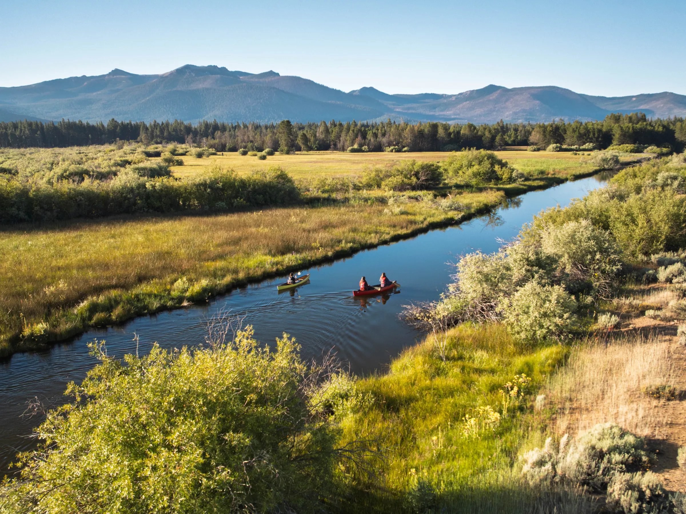 Two people canoe on a river surrounded by grassy fields and mountains in the background.