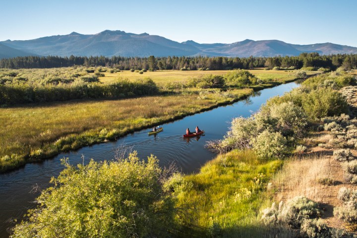 Two people canoe on a river surrounded by grassy fields and mountains in the background.