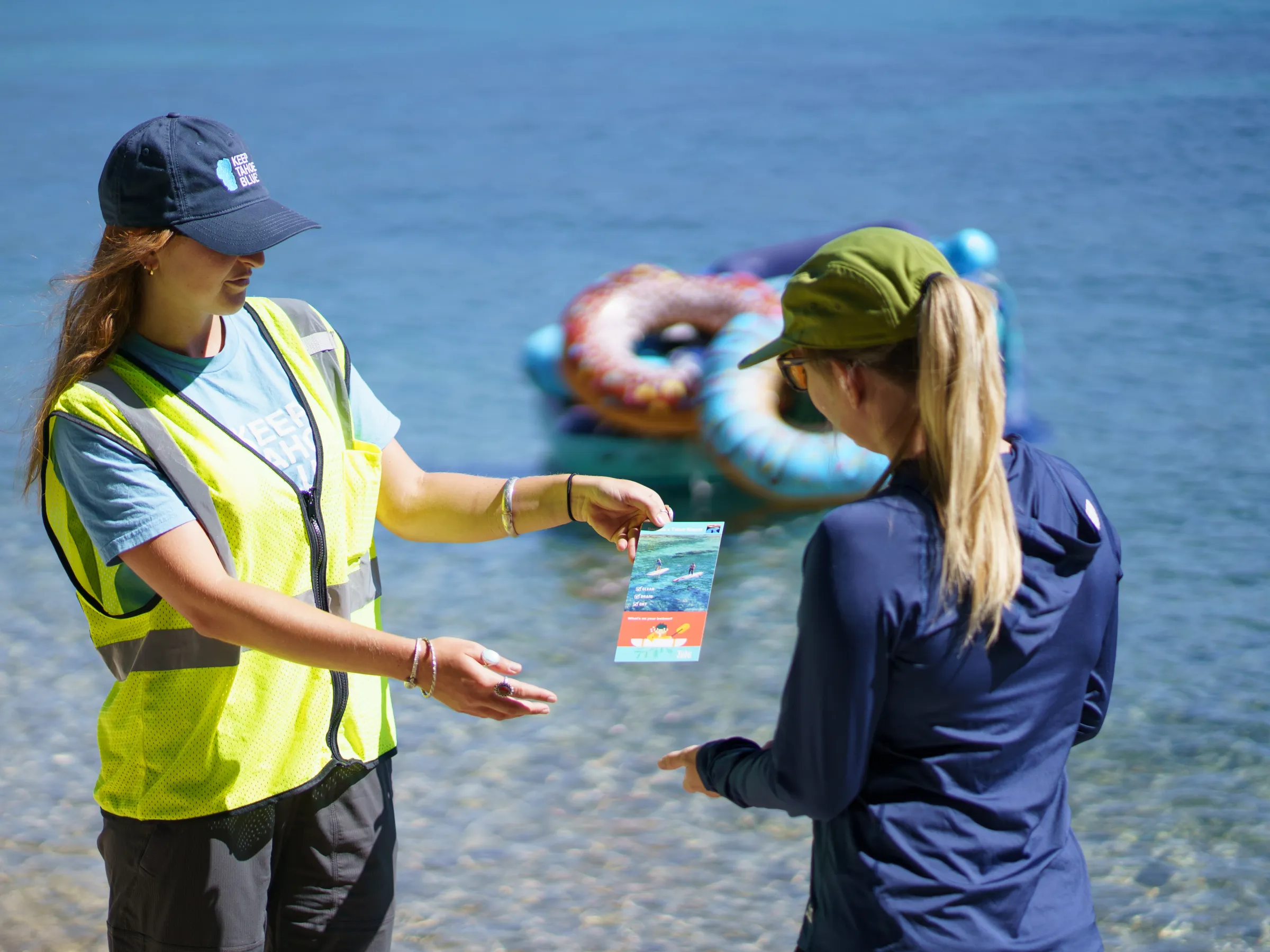 Two women exchanging pamphlet by the beach, with inflatable rings in background.