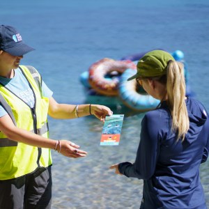 Two women exchanging pamphlet by the beach, with inflatable rings in background.