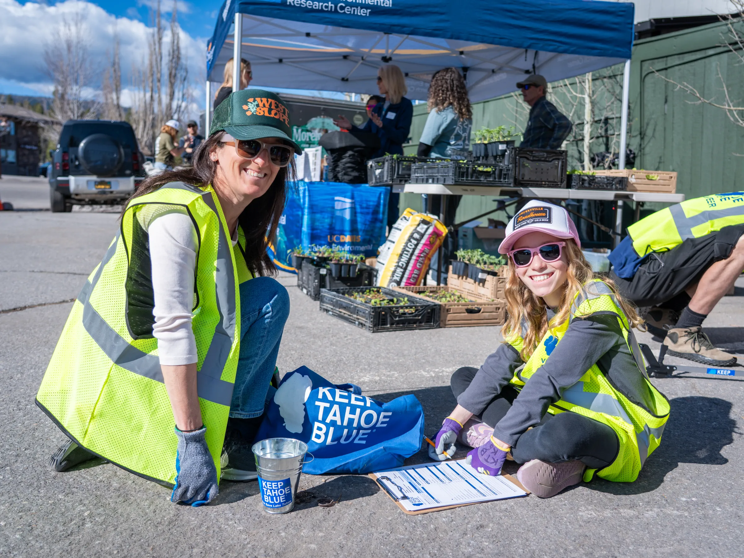Two women in vests planting at an outdoor event with a 'Keep Tahoe Blue' sign.