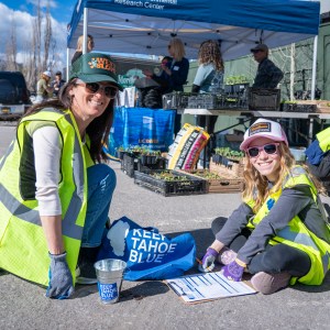 Two women in vests planting at an outdoor event with a 'Keep Tahoe Blue' sign.