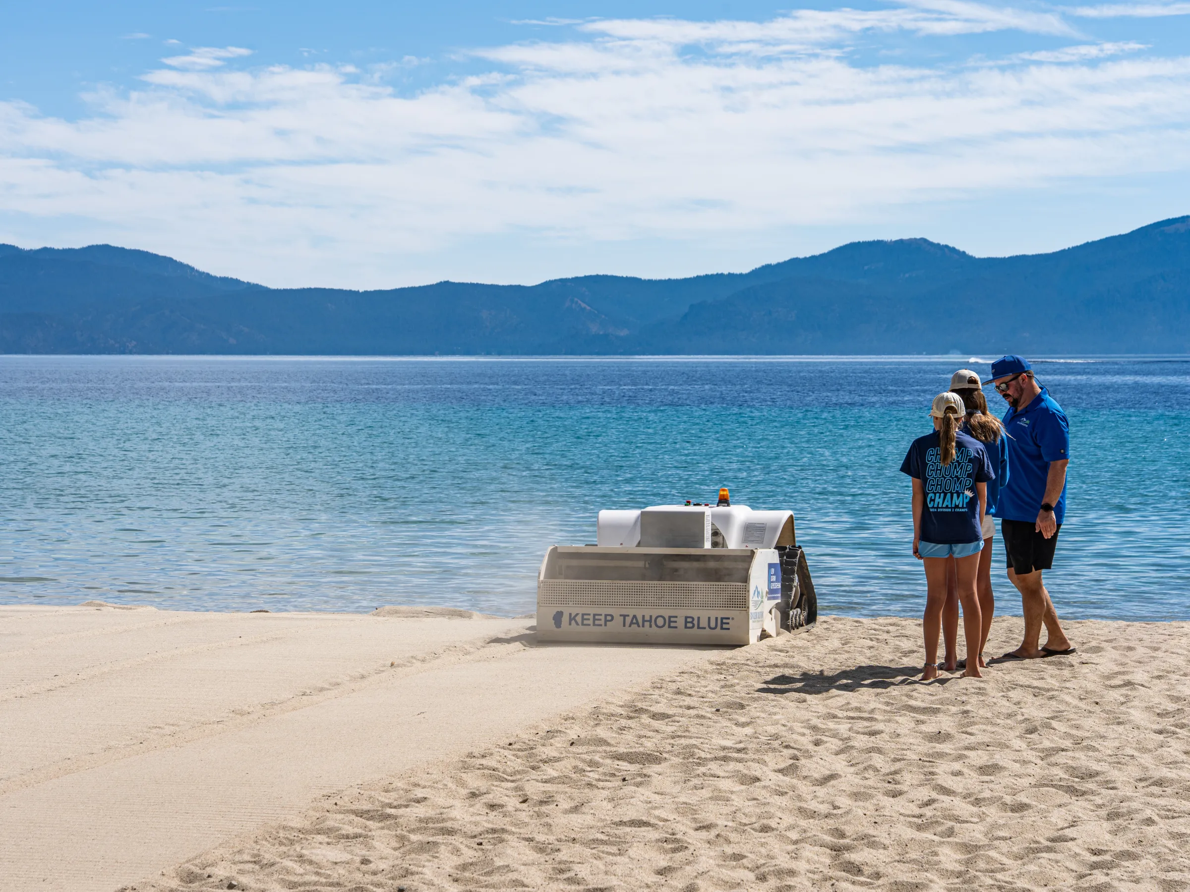People stand near a sandy beach with a 'Keep Tahoe Blue' cleaner and scenic mountains in the background.