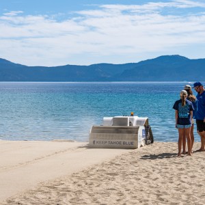 People stand near a sandy beach with a 'Keep Tahoe Blue' cleaner and scenic mountains in the background.