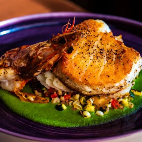 Grilled fish and shrimp on green puree with diced vegetables on a plate.