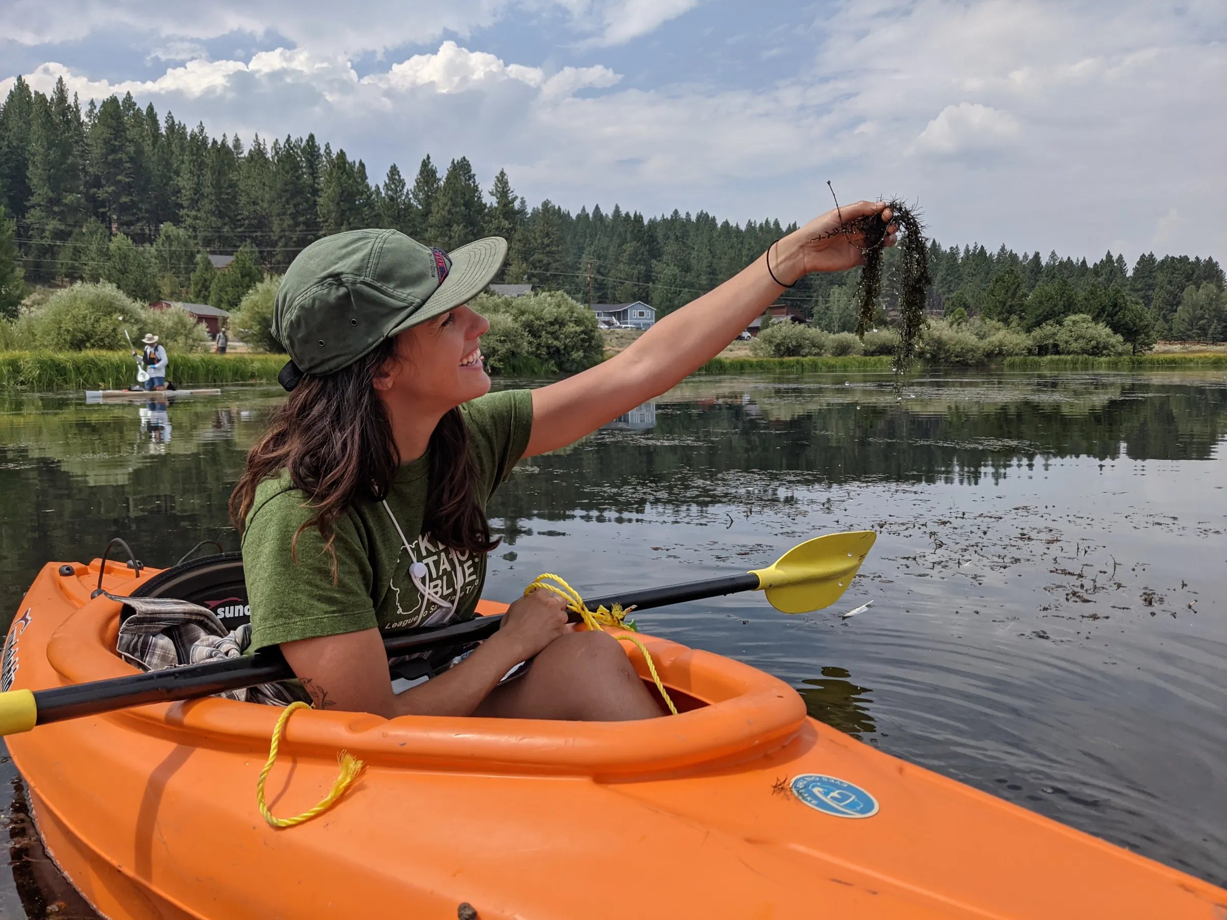 Woman in orange kayak holding seaweed on a lake with trees in background.