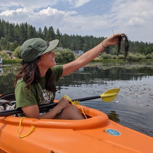 Woman in orange kayak holding seaweed on a lake with trees in background.