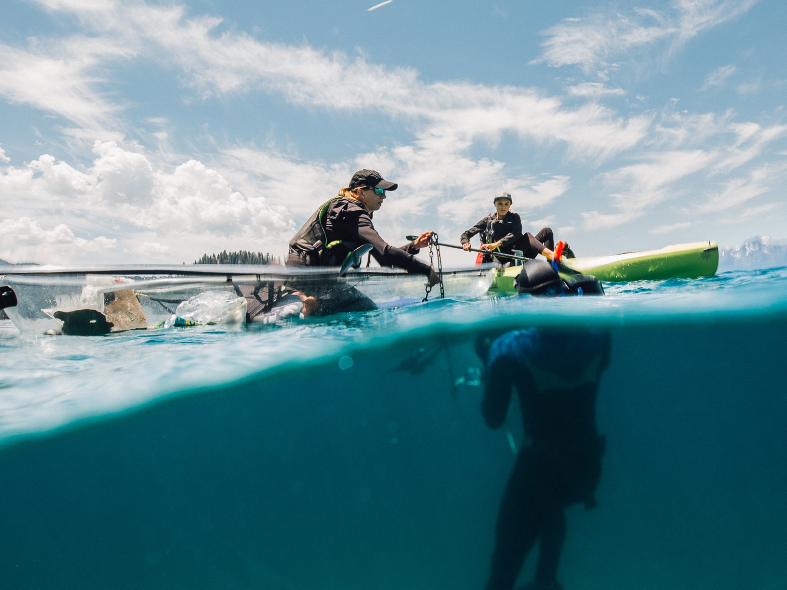 Divers near surface next to kayak in clear water, mountains in background.