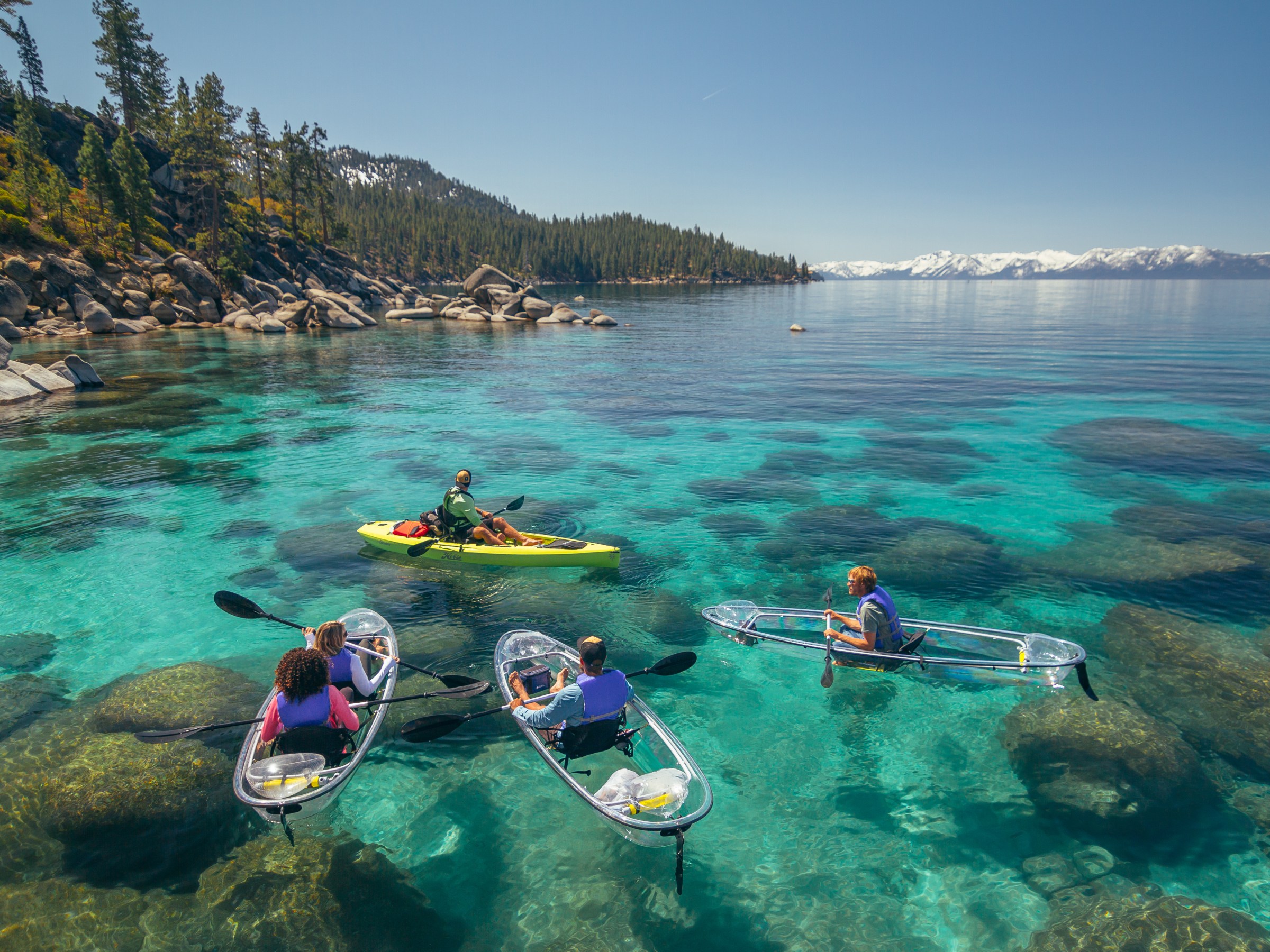 People kayaking on clear, turquoise water with rocky shoreline and snowy mountains in the background.