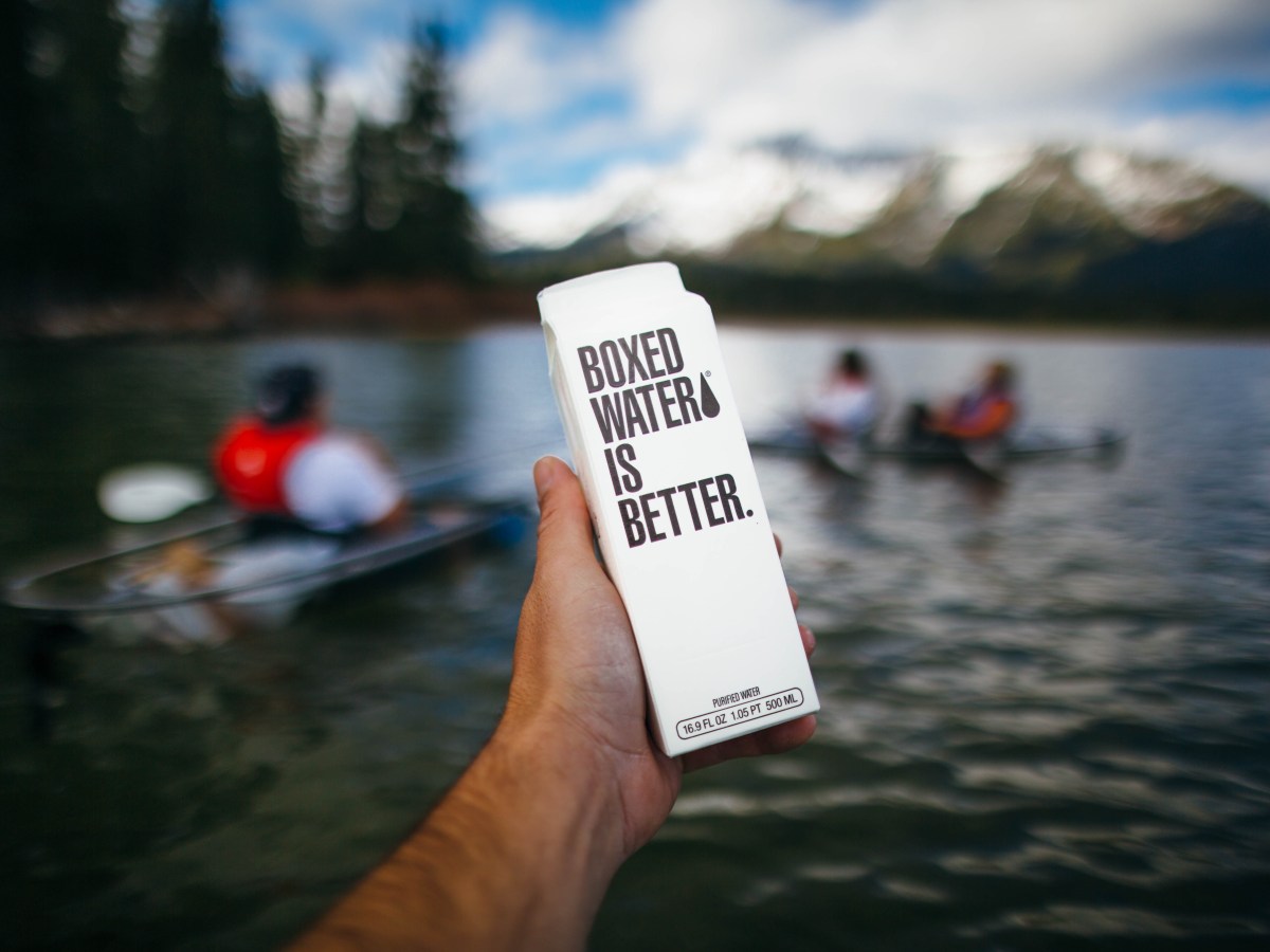 Hand holding boxed water with lake and kayakers in the background.