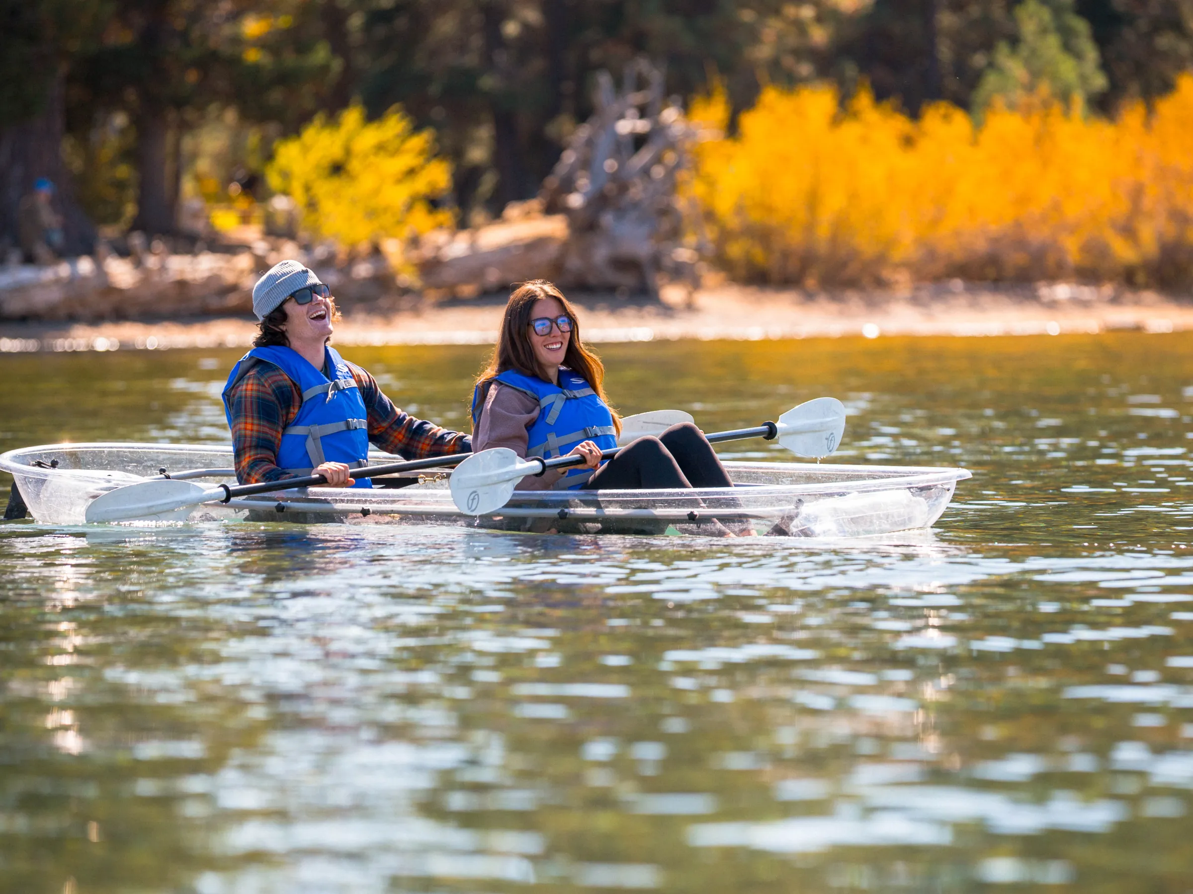 Two people in life vests kayaking on a clear boat in a lake with autumn trees in the background.