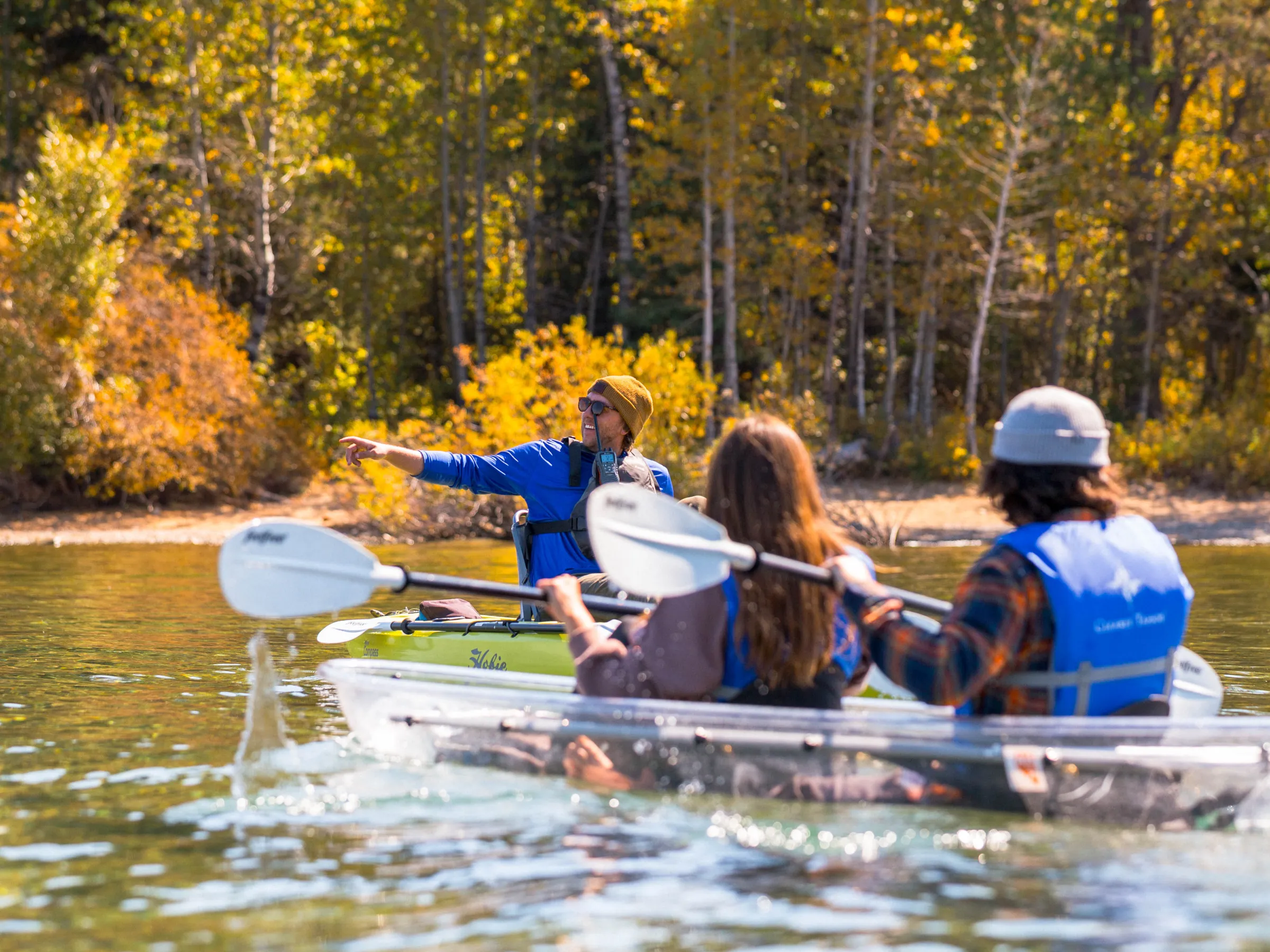 Three people kayaking on a lake surrounded by autumn trees.