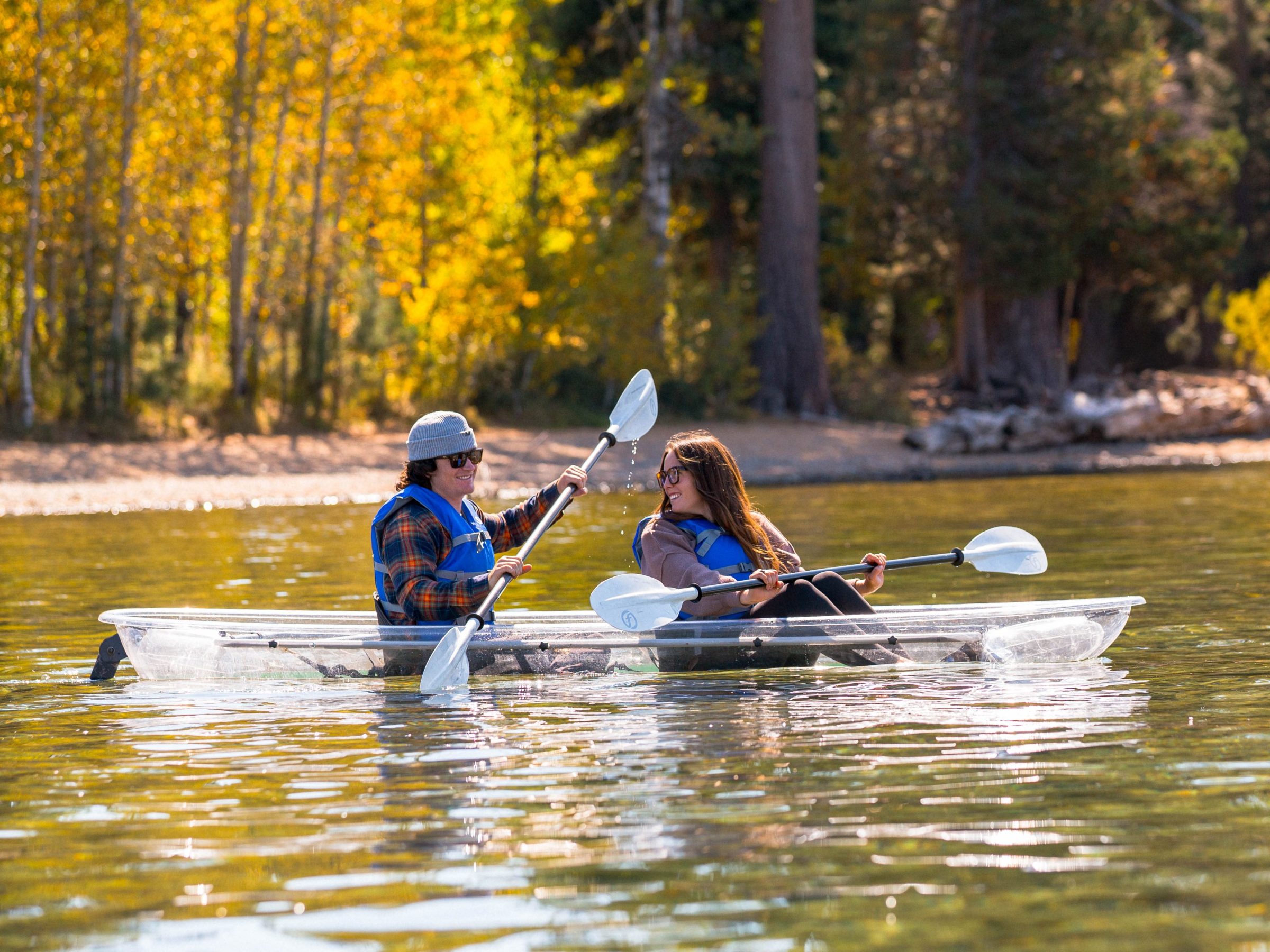 Two people kayaking in clear waters near a forest in autumn.