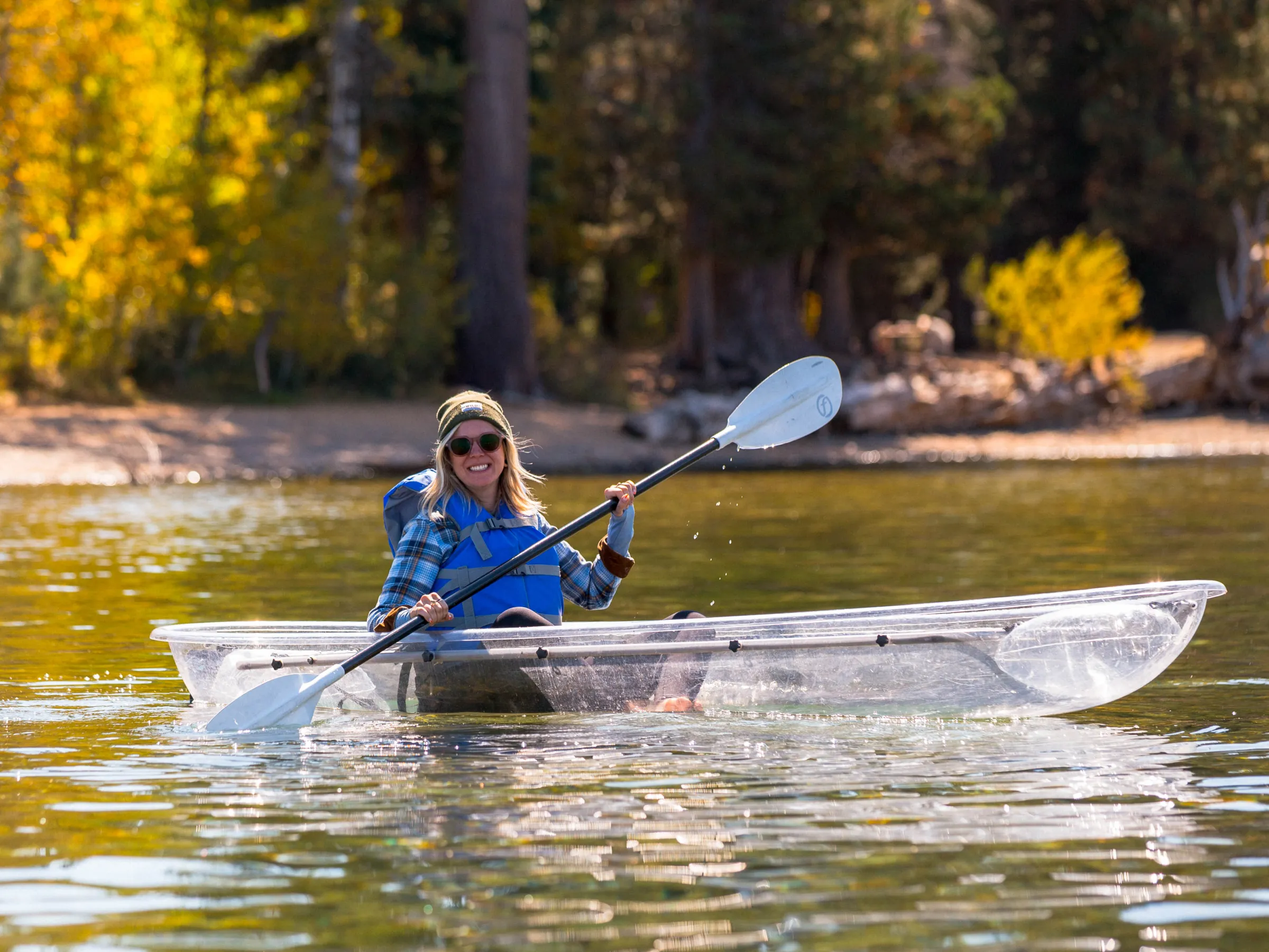 Person kayaking in clear kayak on calm lake with trees in background.