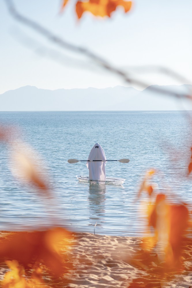 Person in ghost costume kayaking on a lake, framed by autumn leaves.